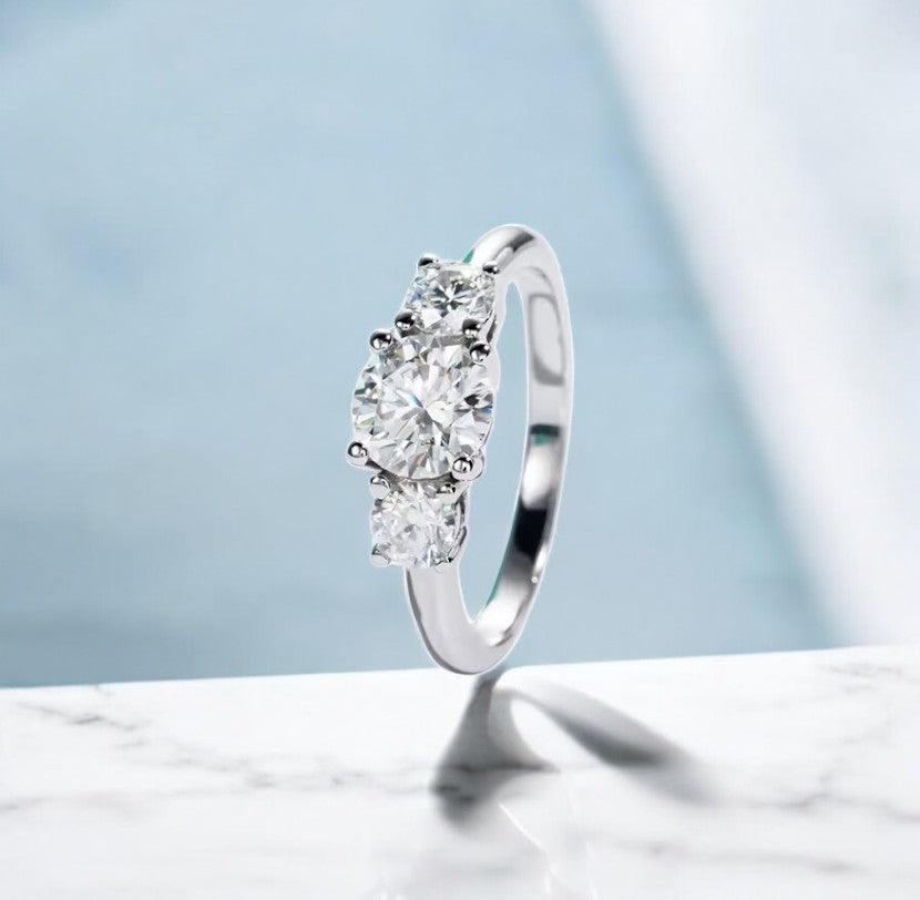 Moissanite engagement ring with 3 stone design, 1 carat main stone and 925 sterling silver, with D color and VVS clarity, on a marble countertop, made by russmoon